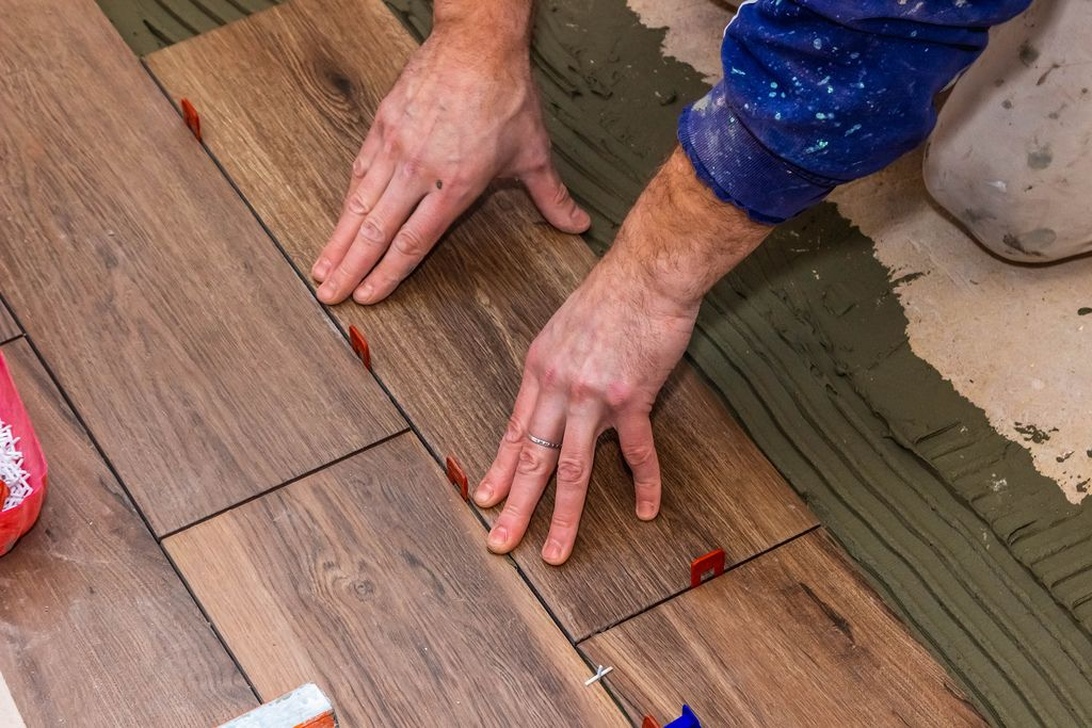 Närbild av händer som installerar klinkerplattor med träutseende. Installatören placerar och justerar plattor på ett förberett murbruk med röda kakelkryss synliga mellan plattorna.
